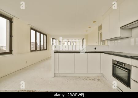 Das Innere der leeren offenen Küche mit weißen Schränken und schwarzer Theke in der neuen Wohnung mit großen Fenstern und weißen Wänden Stockfoto