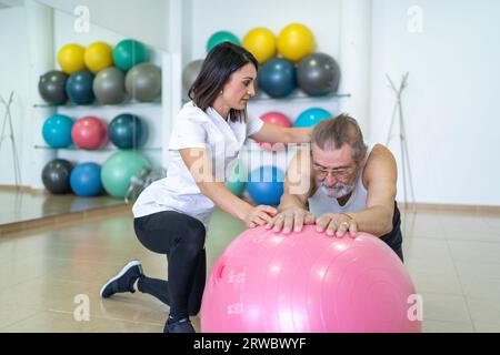 Konzentrierte Ärztin bei Uniform-assistierenden älteren männlichen Patienten bei körperlicher Bewegung am Fitnessball während des Rehabilitationsverfahrens unter Verachtung Stockfoto