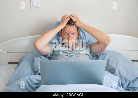 Gut aussehender beeindruckter Mann, der am Morgen Nachrichten auf dem Laptop las, mit verärgertem Ausdruck im Bett. Der Mann staunt über schlechte Nachrichten, die er am Computer ansieht, hält einen Kopf Stockfoto