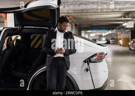 Mann mit Mobilgerät, der das Laden von Elektrofahrzeugen im Untergeschoss beobachtet Stockfoto