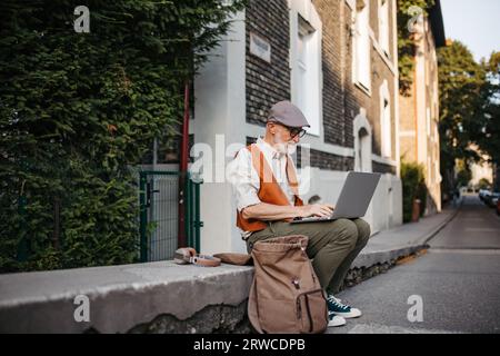 Älterer Mann sitzt auf einer Bordsteinkante und arbeitet an seinem Laptop. Stockfoto