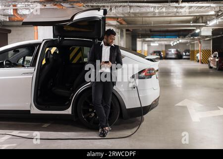 Mann mit Mobilgerät, der das Laden von Elektrofahrzeugen im Untergeschoss beobachtet Stockfoto