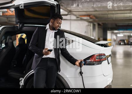 Geschäftsmann, der das Elektrofahrzeug über die mobile App auf dem Parkplatz im Untergeschoss auflädt Stockfoto