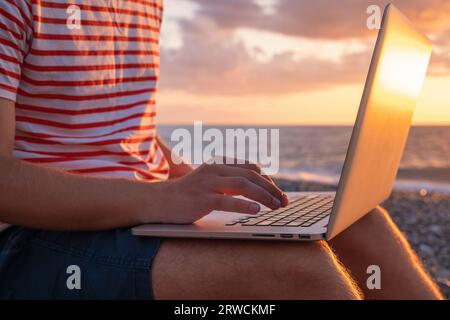 Nahaufnahme eines digitalen Nomaden, der mit einem Laptop arbeitet, sitzt am Strand am Meer während des Sonnenuntergangs. Männliche Freiberufler arbeiten aus der Ferne online. Remote-Arbeit Stockfoto