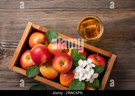 Reife rote Äpfel in Holzkiste mit Zweig weißer Blumen und einem Glas frischen Saft auf einem Holztisch. Draufsicht. Stockfoto