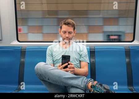 Junger kaukasischer Mann, der in der U-Bahn im Zug sitzt und Handy benutzt. Männlicher Reisender im U-Bahn-Zug, der im Internet surft und soziale Medien anschaut Stockfoto