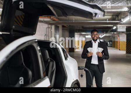 Ein Geschäftsmann hält Banknoten in der Nähe der Parkgarage, um ein E-Auto zu laden Stockfoto