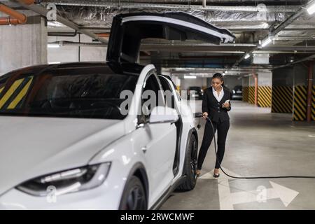 Dame mit Smartphone, die EV-Ladegerät in der öffentlichen Garage anschließt Stockfoto