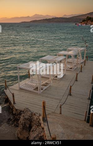 Vertikaler Blick auf den hölzernen Pier mit Liege während der Goldenen Stunde in Ksamil. Wunderschöne albanische Riviera mit Sonnenuntergangsfarben. Ionisches Meer und Berge. Stockfoto