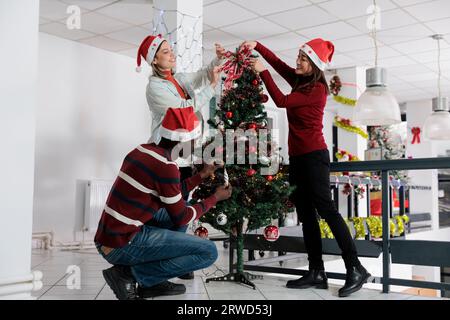 Mitarbeiter, die den Weihnachtsbaum mit Ornamenten und Lichtergirlanden dekorieren, arbeiten während der Winterferien. Mitarbeiter, die gemeinsam im festlichen Büro verbringen Stockfoto