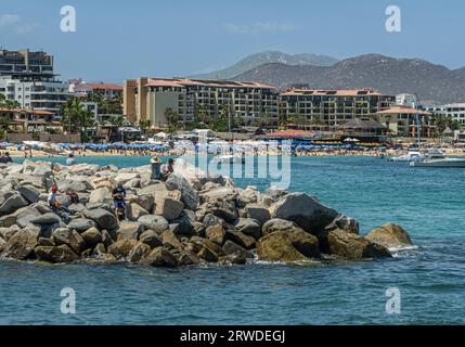 Mexiko, Cabo San Lucas - 16. Juli 2023: Fin del Muelle felsige Halbinsel am Hafenausgang mit Hacienda Strand in der Rückseite voll von blauen Sonnenschirmen und Wand von Tal Stockfoto