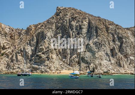 Mexiko, Cabo San Lucas - 16. Juli 2023: Kleiner Strand, viele Badende, unter hohen grauen felsigen Klippen in der Nähe von Landsend mit mehreren kleinen Booten auf grünem Ba Stockfoto