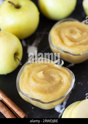 Hausgemachtes Bio-Apfelpüree mit einem Drall auf der Oberseite in einer Glasschale mit Zimt und goldenem köstlichem Apfel auf dunklem Hintergrund. Gemischte Babynahrung. Stockfoto