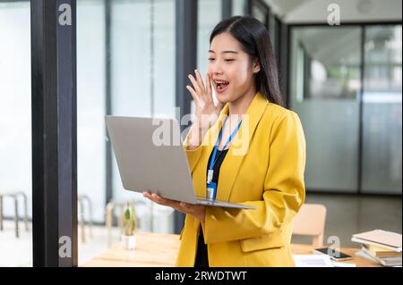 Eine fröhliche und aufgeregte asiatische Geschäftsfrau im gelben Anzug, die in ihrem Büro auf ihren Laptop-Bildschirm schaut, während sie mit einem aufgeregten Gesicht steht. Stockfoto