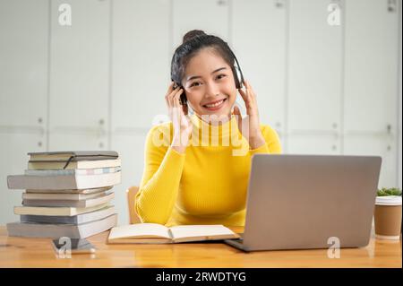 Ein Porträt einer attraktiven und lächelnden jungen asiatischen Universitätsstudentin sitzt mit ihrem Laptop und einem Stapel Bücher an einem Tisch in einer Bibliothek. Stockfoto