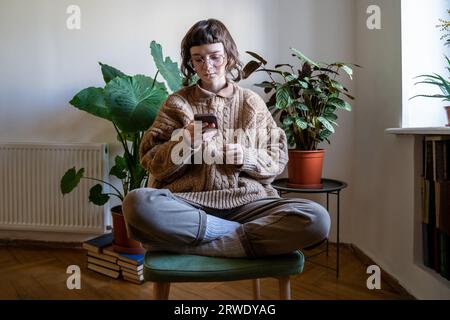 Das Mädchen benutzt das Telefon und sitzt auf dem Stuhl. Frau hält Smartphone, scrollt durch die sozialen Netzwerke der Nachrichtenfeeds Stockfoto