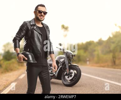 Biker in einer Lederjacke mit geparktem Motorrad auf der Straße, Helm haltend und Sonnenbrille tragend Stockfoto
