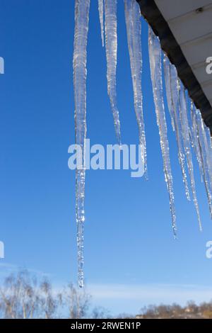 Eiszapfen hängen auf dem Dach eines Hauses vor einem hellblauen Himmel. Frühlingslandschaft mit Eiszapfen, die vom Dach des Hauses hängen. Schneeeis, Stockfoto