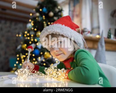 Das Weihnachtskind schreibt einen Brief an den Weihnachtsmann, während es am Tisch vor einem Bokeh-Hintergrund sitzt. 7-jähriger Junge in rotem Weihnachtsmannhut träumt von Geschenken zu Hause. Gif Stockfoto