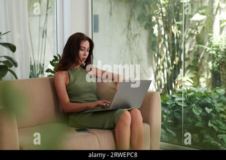 Junge elegante Geschäftsfrau, die zu Hause auf dem Sofa sitzt und am Laptop arbeitet Stockfoto