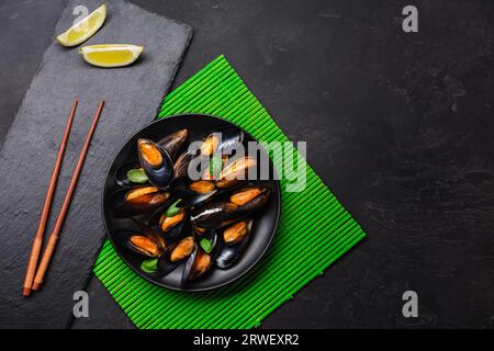 Muscheln aus Meeresfrüchten, Basilikumblätter auf einem schwarzen Teller mit Zitrone und Stäbchen auf grüner Bambusmatte und Steintisch. Draufsicht. Stockfoto