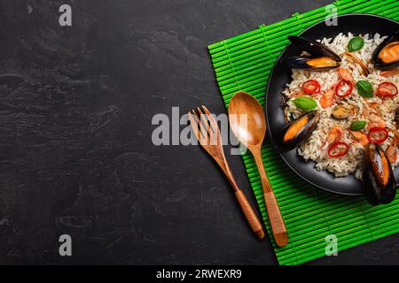 Gebratener Reis mit Muscheln, Garnelen, Basilikum auf schwarzem Teller mit Holzlöffel und Gabel auf grüner Bambusmatte und Steintisch. Draufsicht. Stockfoto