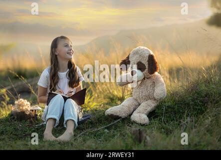 Junge, lächelnde Frau, die bei Sonnenuntergang in der Natur sitzt, mit ihrem Teddy, der in ihr Buch schreibt Stockfoto