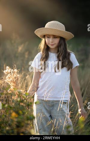 Das junge Mädchen, lässig gekleidet und mit einem Hut, steht auf einem Feld und hält ein Stück getrocknetes Gras Stockfoto