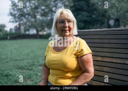Glückliche Seniorin, die sich auf der Bank im Rasen entspannt. Nahgesicht einer Reifen blonden Frau, die lächelt und in die Kamera schaut. Pensionierte Frau in Gelegenheitsfällen Stockfoto