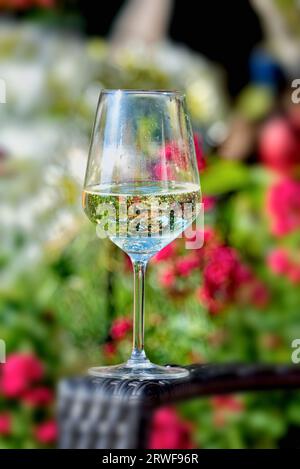 Nahaufnahme eines Glases Weißwein auf dem Arm eines Gartenstuhls mit einem weichen Fokus auf bunten Gartenpflanzen im Hintergrund an einem Sommertag Stockfoto