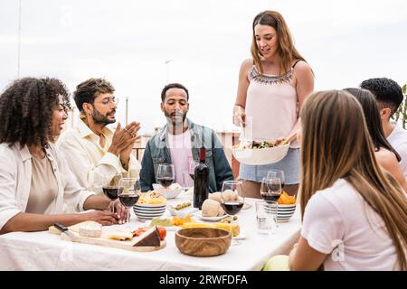 Junge Frau, die ihren Freunden Salat auf einer Dinnerparty serviert. Eine Gruppe von Freunden verschiedener Rassen, die zusammen auf der Dachterrasse Dinner feiern. Stockfoto