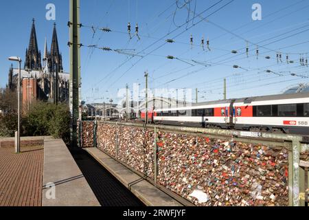 Der Zug verlässt den Kölner Bahnhof, mit einem Zaun, der im Vordergrund mit „Liebesschloss“-Vorhängeschlössern versehen ist, und dem Dom auf der linken Seite. Stockfoto