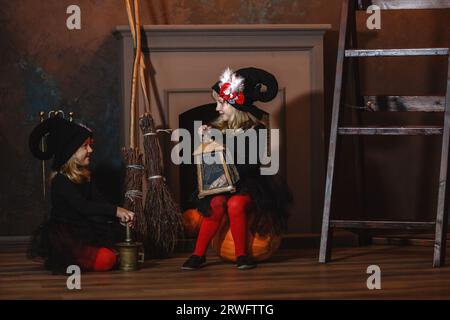 Mädchen in Hexenkostümen sitzen am Kamin auf Kürbissen, eine hält eine Laterne, die andere macht einen Trank. Halloween. Stockfoto