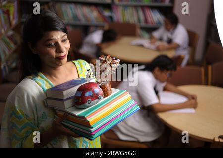 Indische Lehrerin hält ein Bündel Bücher in der Hand, steht in der Bibliothek, studiert Druck, erzieherische Kraft. Stockfoto