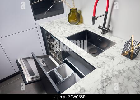 Küche mit eingebautem, geöffnetem Geschirrspüler und herausziehbarer Schublade für die getrennte Abfallsammlung. Wasserhahn Waschbecken mit kompaktem Hochdruck Stockfoto