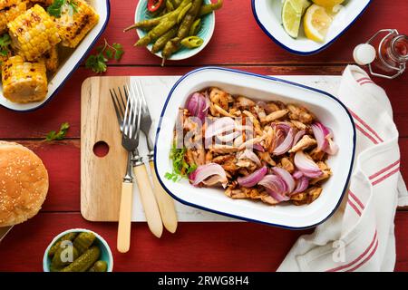 Gezapftes bbq-Hühnchen mit gebackenen Zwiebeln auf Servierplatte, bbq-Mais, Gurken, Chilischoten und Brötchen für Hot Dogs und Burger, Tomatensauce. Traditionell Stockfoto