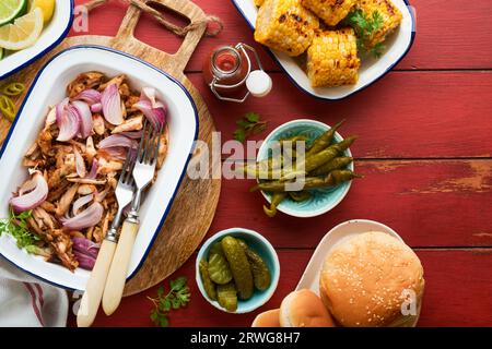 Gezapftes bbq-Hühnchen mit gebackenen Zwiebeln auf Servierplatte, bbq-Mais, Gurken, Chilischoten und Brötchen für Hot Dogs und Burger, Tomatensauce. Traditionell Stockfoto