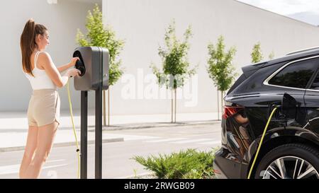 Lächelndes Mädchen, das das Ladegerät in ihr schwarzes Elektroauto steckte und es mit der auf dem Parkplatz installierten EV-Ladestation verband Stockfoto