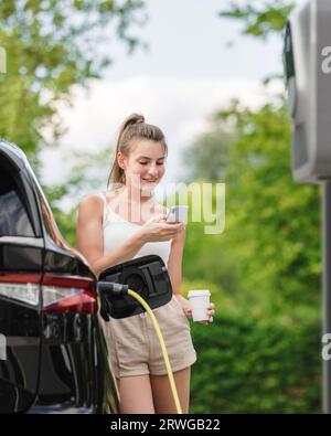 Schönes Mädchen, das ein Kabel-Ladegerät in ihr geparktes schwarzes Elektroauto steckte und ein Smartphone benutzte. Konzept der öffentlichen Ladestation für Elektrofahrzeuge. Stockfoto