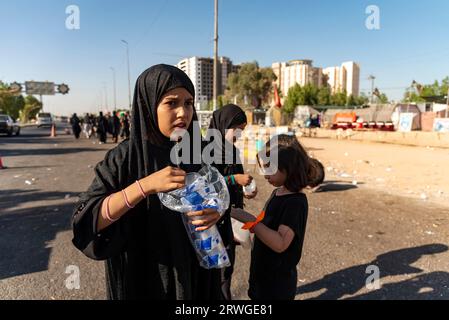 Najaf, Irak. September 2023. Ein irakisches schiitisches Mädchen verteilt Saft unter schiitischen Pilgern, die von Najaf in Richtung der Heiligstadt Karbala marschieren. Jedes Jahr pilgern Millionen schiitische Muslime und einige aus anderen Glaubensrichtungen zu Fuß von verschiedenen Städten im Irak und Iran in die heilige Stadt Karbala. Diese Pilgerfahrt erinnert an Imam Hussein, den Enkel des Propheten Mohammed, der 680 n. Chr. in einer Schlacht starb. Am 40. Tag der Trauer um Hussein, bekannt als Arbaeen, kommen Pilger in Karbala zusammen, um an seinem Heiligtum Tribut zu zollen. Auf dem Weg stellen Freiwillige Nahrung, Wasser, Stockfoto