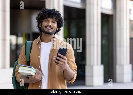 Porträt eines jungen indischen Studenten mit Rucksack, der Telefon und Bücher hält und lächelnd auf die Kamera schaut, während er auf dem Universitätscampus steht. Stockfoto