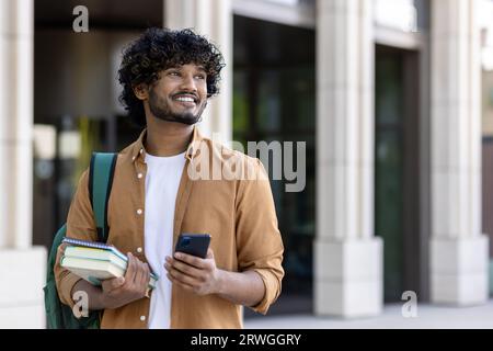 Lächelnder junger indischer Student, der vor dem Campus steht, Bücher hält, Rucksack trägt und Telefon benutzt. Sieht zur Seite. Stockfoto
