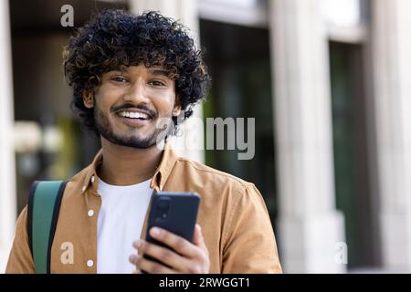 Nahaufnahme eines jungen Indianers, der in der Kamera lächelt und ein Telefon und einen Rucksack hält. Stockfoto