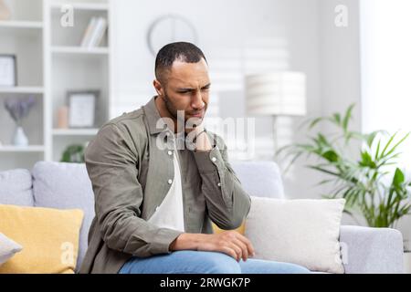 Ein junger muslimischer Mann leidet unter Nackenschmerzen. Er sitzt ermüdend auf der Couch und massiert seine Hand. Stockfoto