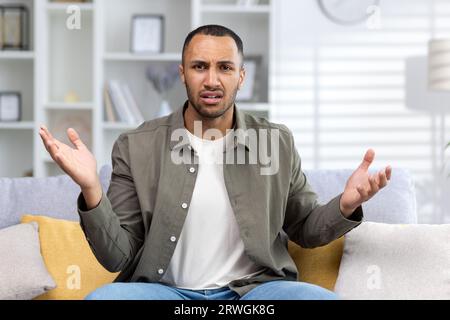 Unzufriedener und verärgerter junger afroamerikanischer Mann sitzt zu Hause auf dem Sofa vor der Kamera, redet, diskutiert. Wirft seine Hände in Frustration auf. Stockfoto