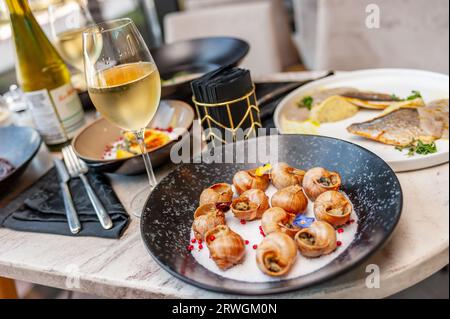 Gekochte Traubenschnecken in Muscheln, mit grünem Öl und Knoblauch auf der Oberseite gefettet, Seitenansicht auf einem Tisch in einem Restaurant mit einem Glas Weißwein Stockfoto