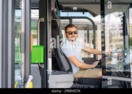 Porträt des Stadtbus-Fahrers mit Sonnenbrille, der in der Fahrerkabine sitzt, während er Passagiere am Busbahnhof wartet. Stockfoto