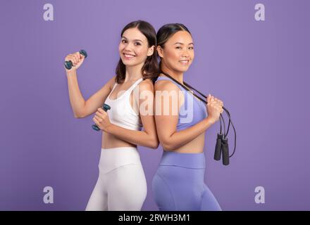Junge asiatische und kaukasische Frauen in Sportkleidung, die mit Hanteln und Sprungseil stehen, violetter Hintergrund Stockfoto