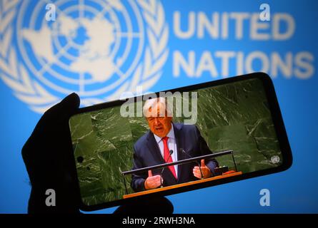 In dieser Abbildung spricht UN-Generalsekretär Antonio Guterres auf der 78. Sitzung der UN-Generalversammlung, die auf einem Smartphone-Bildschirm mit dem Logo der Vereinten Nationen im Hintergrund ausgestrahlt wird. Stockfoto