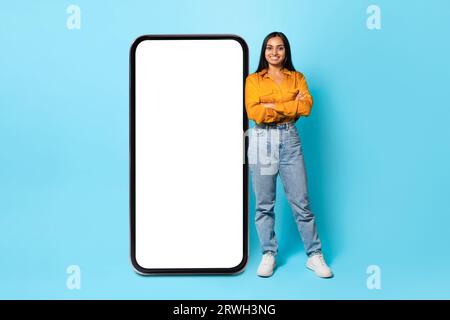 Indische Frau, Die In Der Nähe Des Großen Handybildschirms Auf Blauem Hintergrund Steht Stockfoto
